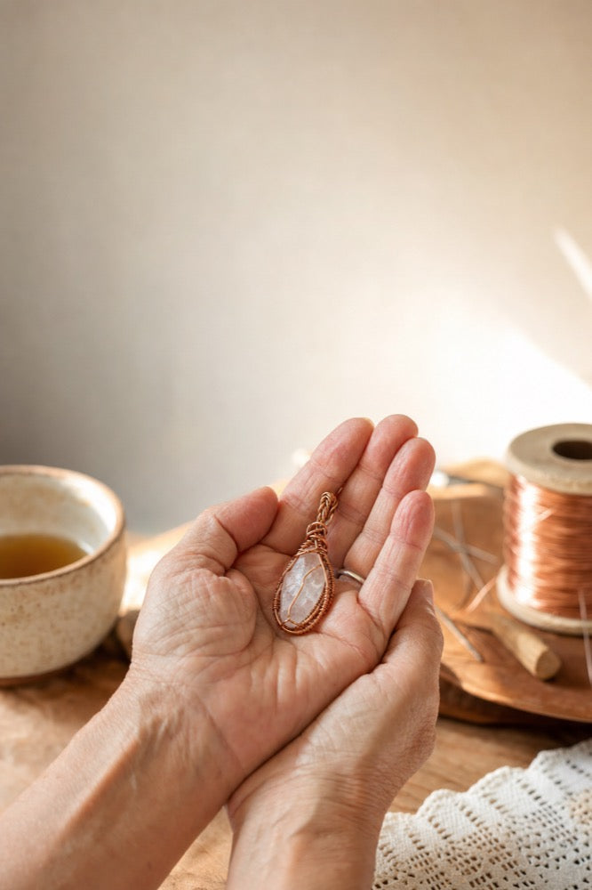 An older woman's hands gently holding a finished wire-wrapped crystal pendant beside a cup of tea